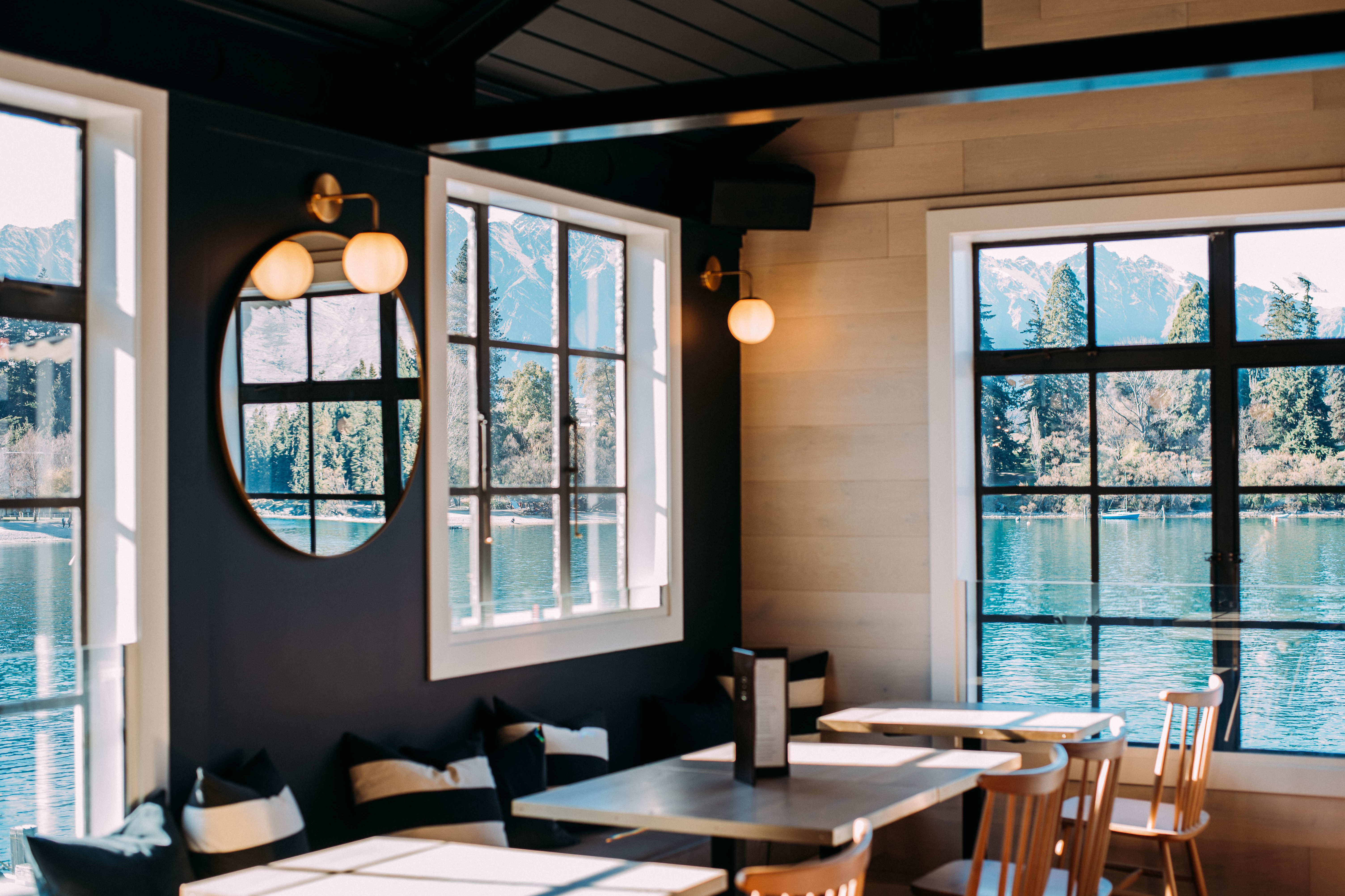 Dark-painted interior with black ceiling beams, round mirror, globe wall lights, and lake and mountain view through white-framed windows