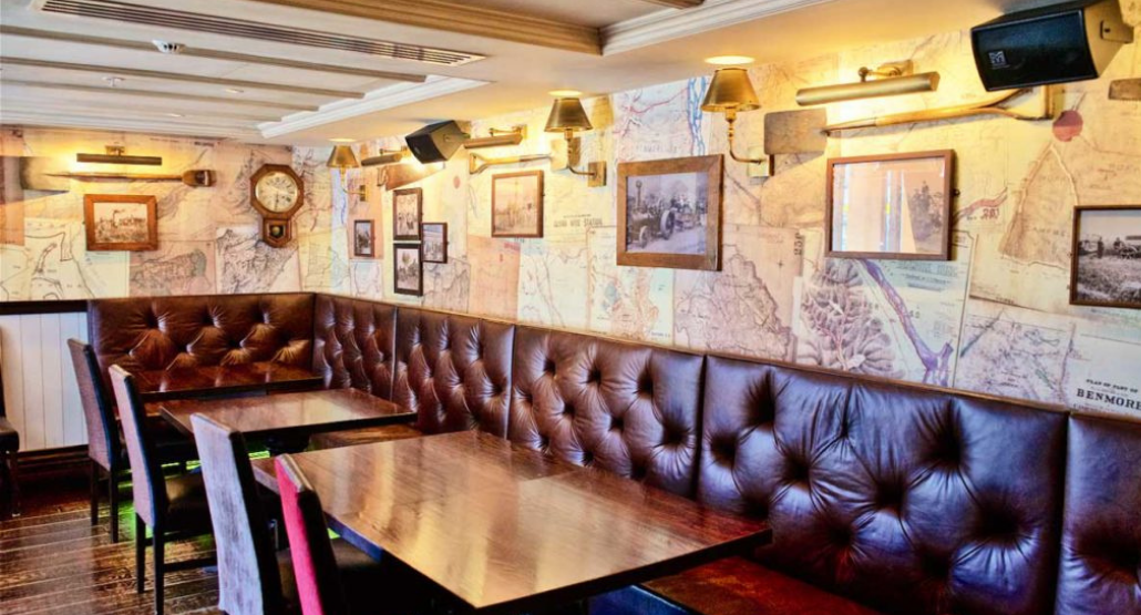 Bar interior with dark timber banquette seating, tufted leather backs, map-print wallpaper, and wall sconce lighting