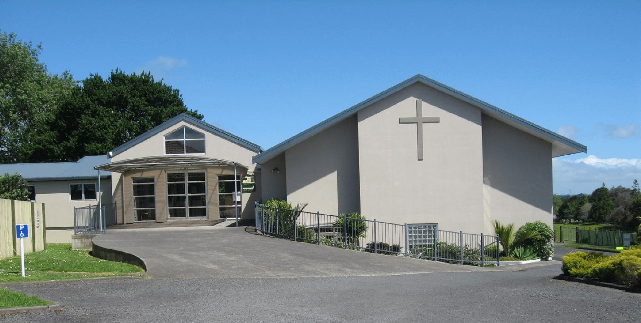 Single-storey church building with cross on gable end, viewed across lawn with hedgerow in foreground