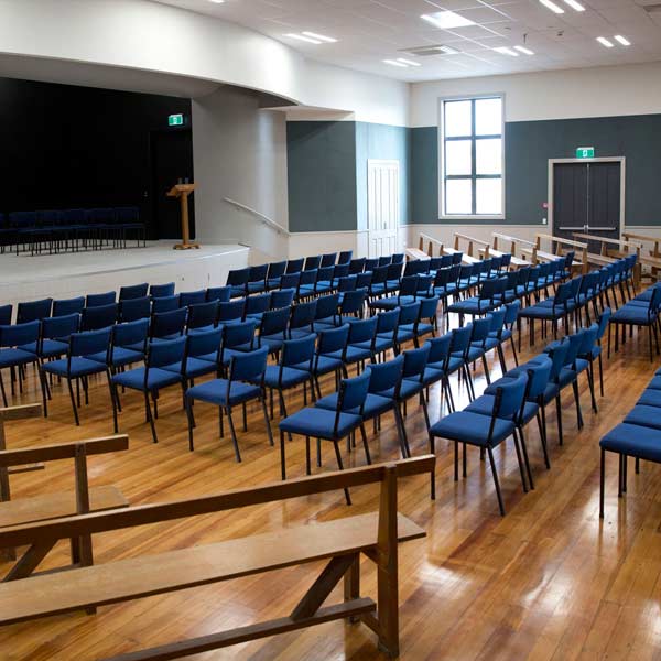 Hall interior with rows of blue chairs facing a raised platform and stage