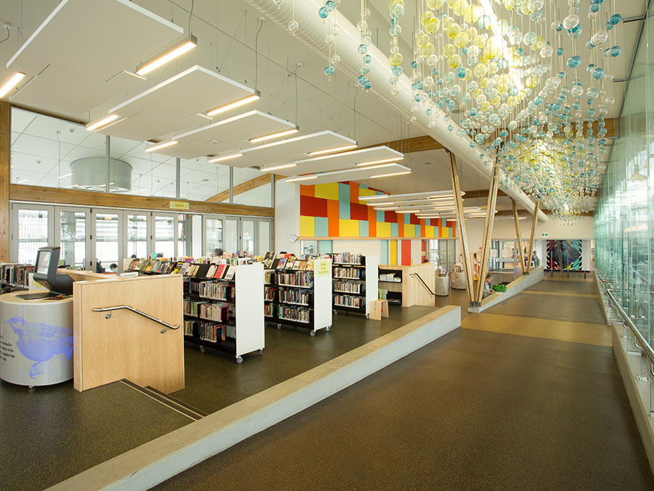 Open-plan library floor with timber shelving, colourful wall panels and glass pendant installation