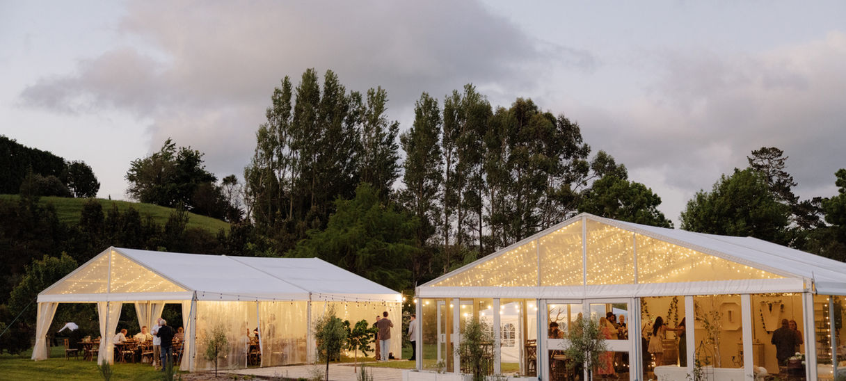 White marquee structures with festoon lights on a hillside at dusk with trees behind