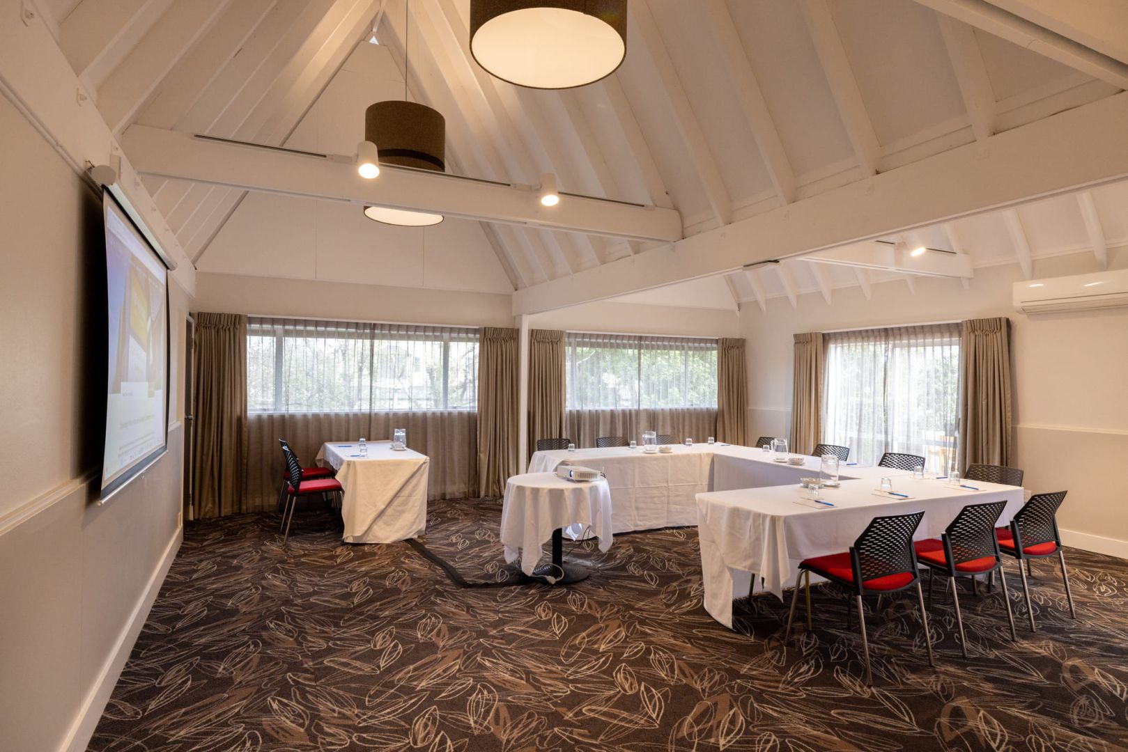 White-plastered function room with vaulted timber-truss ceiling, white tables in U-shape, and large screen