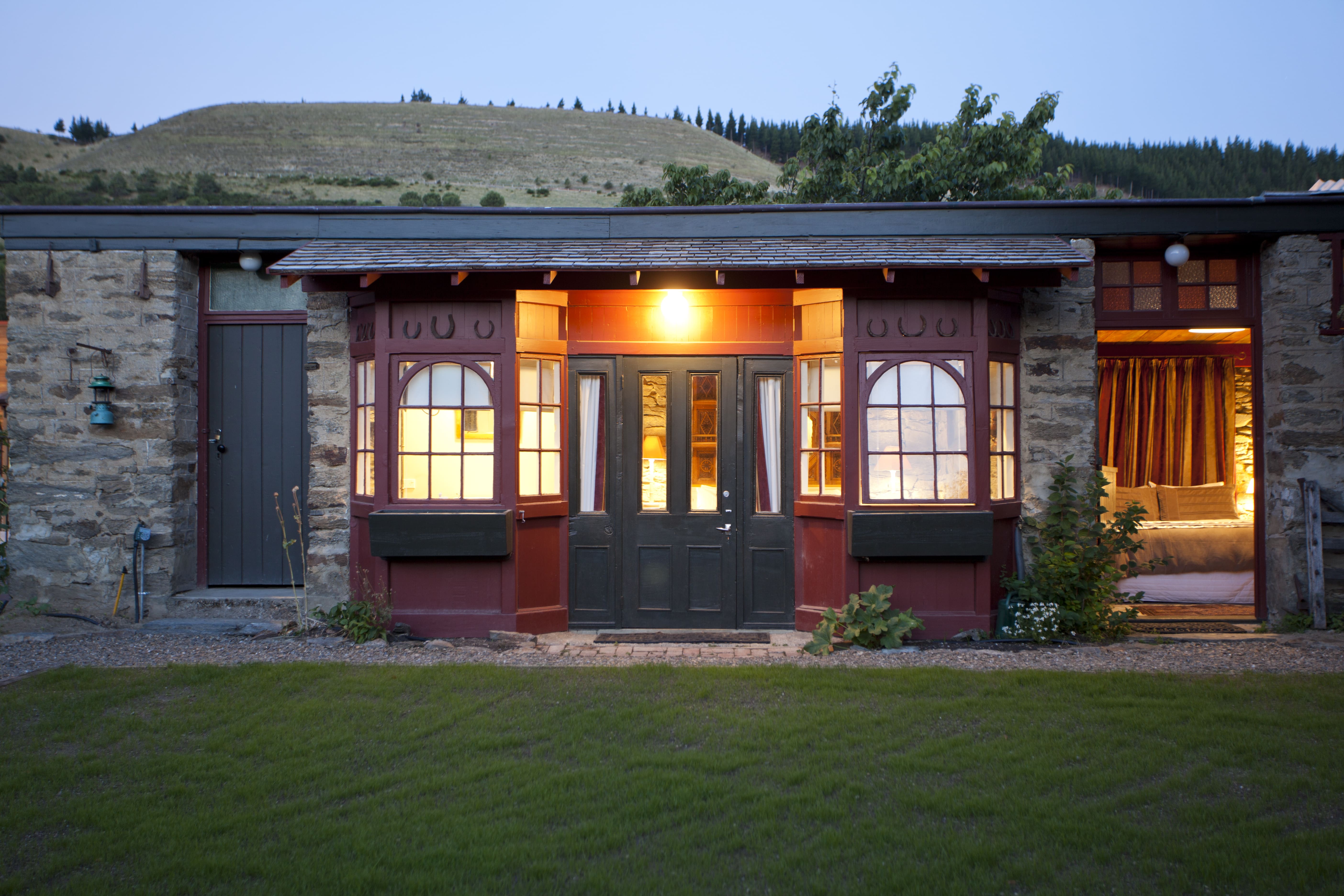 Stone stable building at dusk with lit interiors through arched windows opening onto a lawn