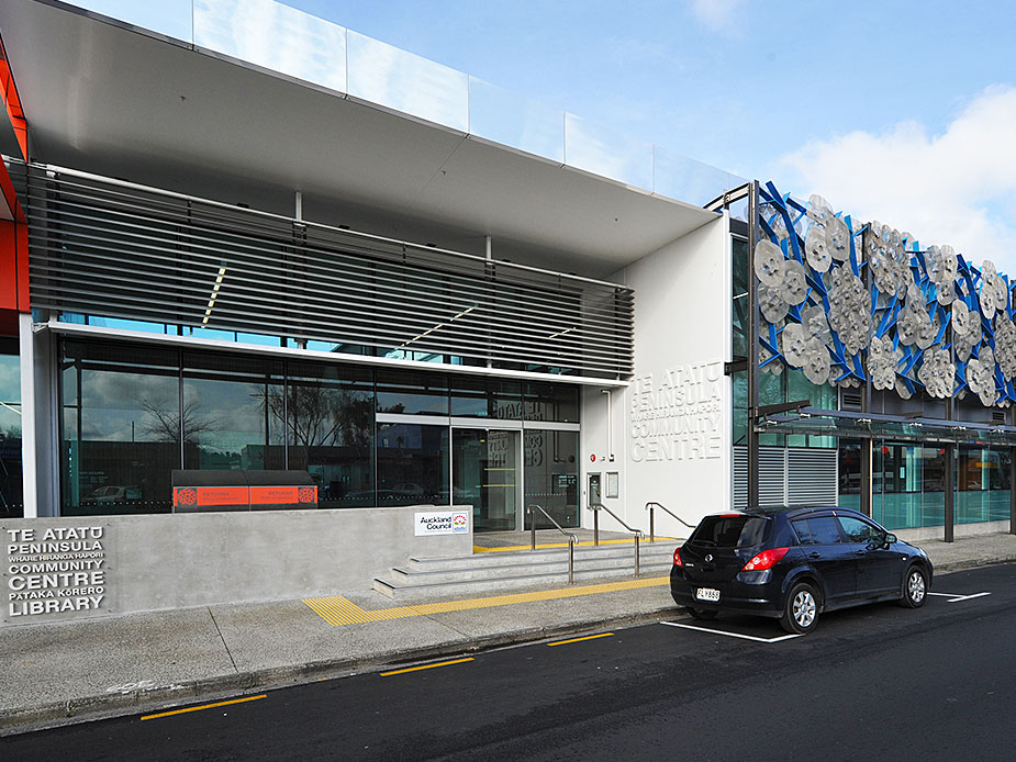 White building facade with louvred sunshade canopy, decorative sculptural panels, and glazed entrance