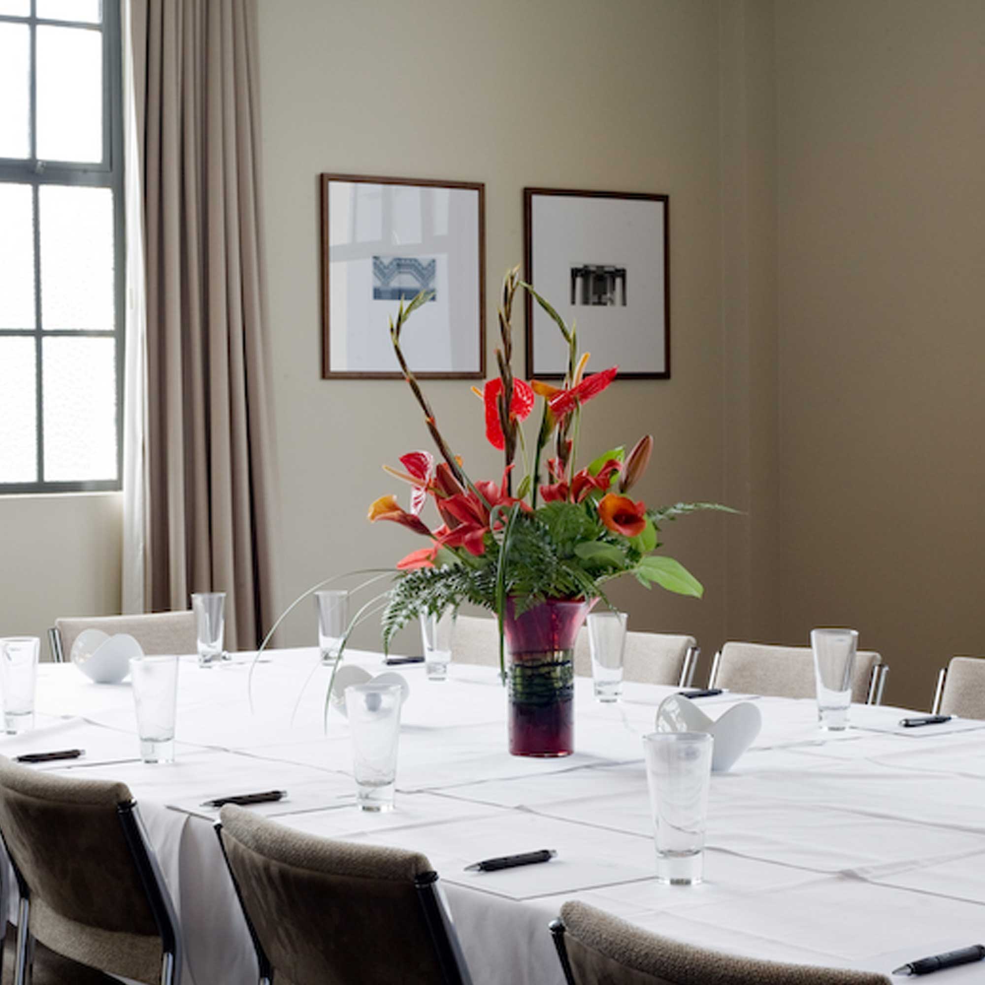Boardroom table with white linen, floral centrepiece, framed prints, large window with curtains