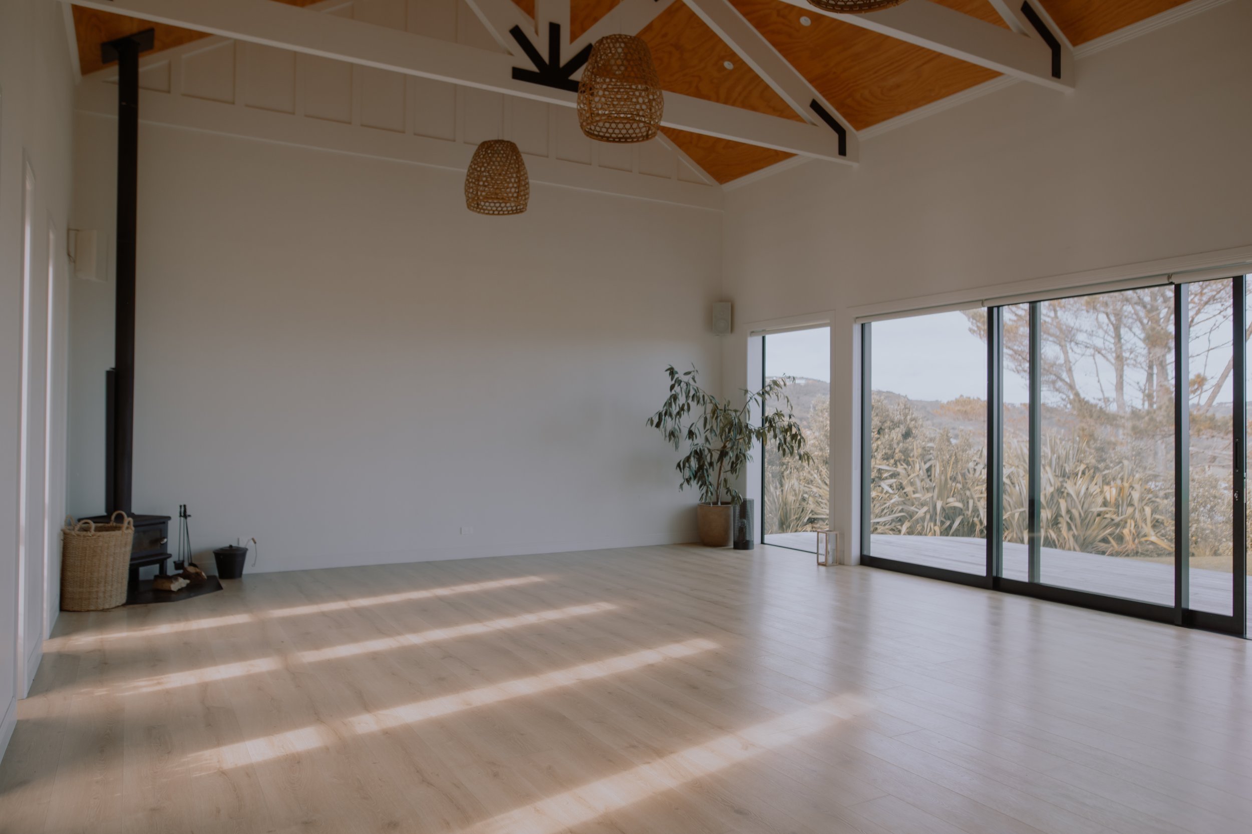 Light-filled open space with vaulted timber ceiling, pale floorboards, wood burner, and sliding glass doors
