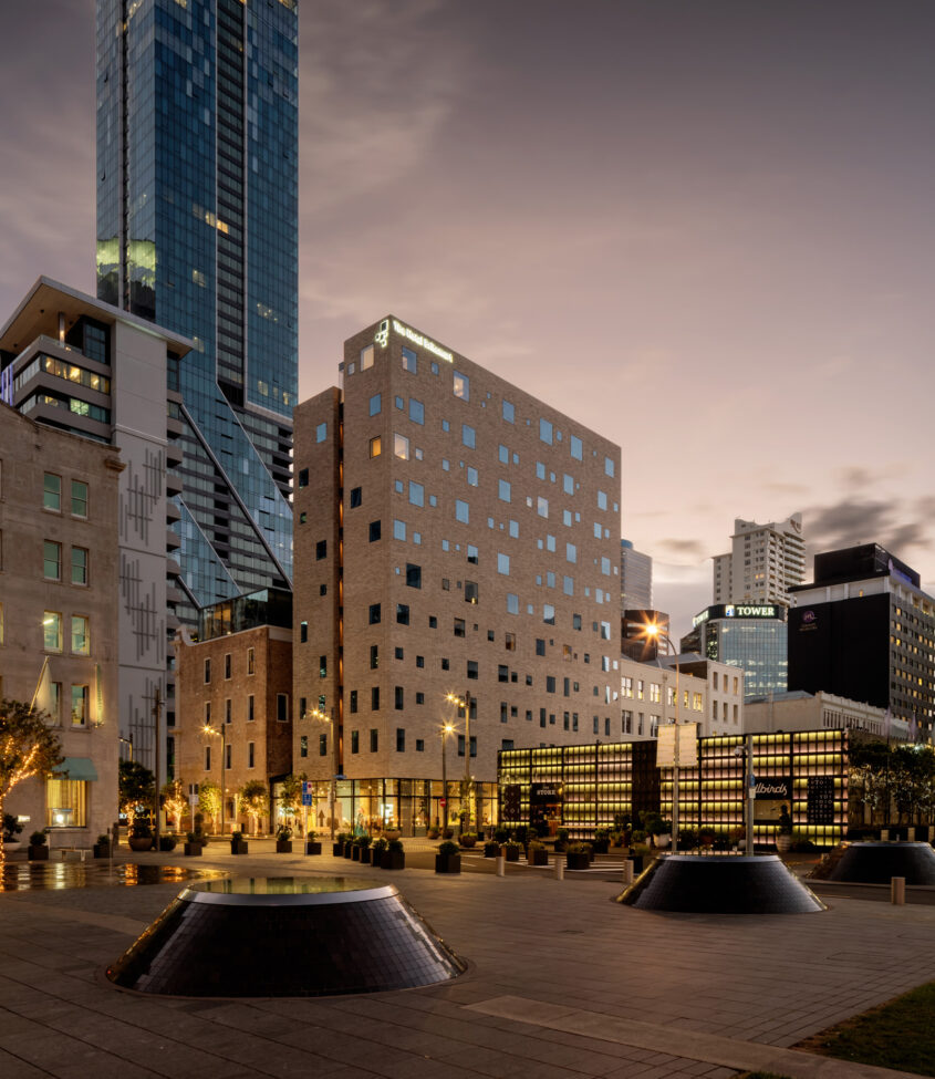 Pale brick multi-storey building with irregular window openings, city skyline at dusk
