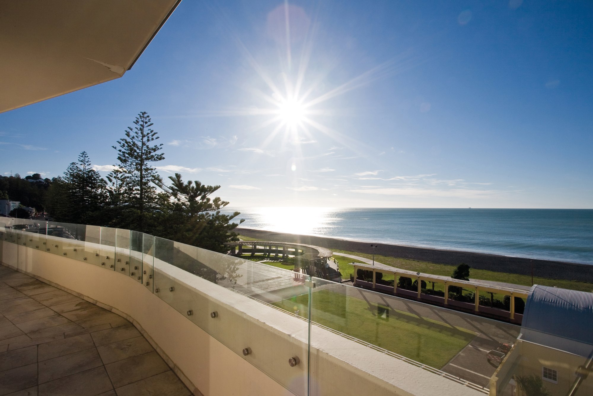 White rendered building balcony with glass balustrade overlooking beach, sea and pine trees