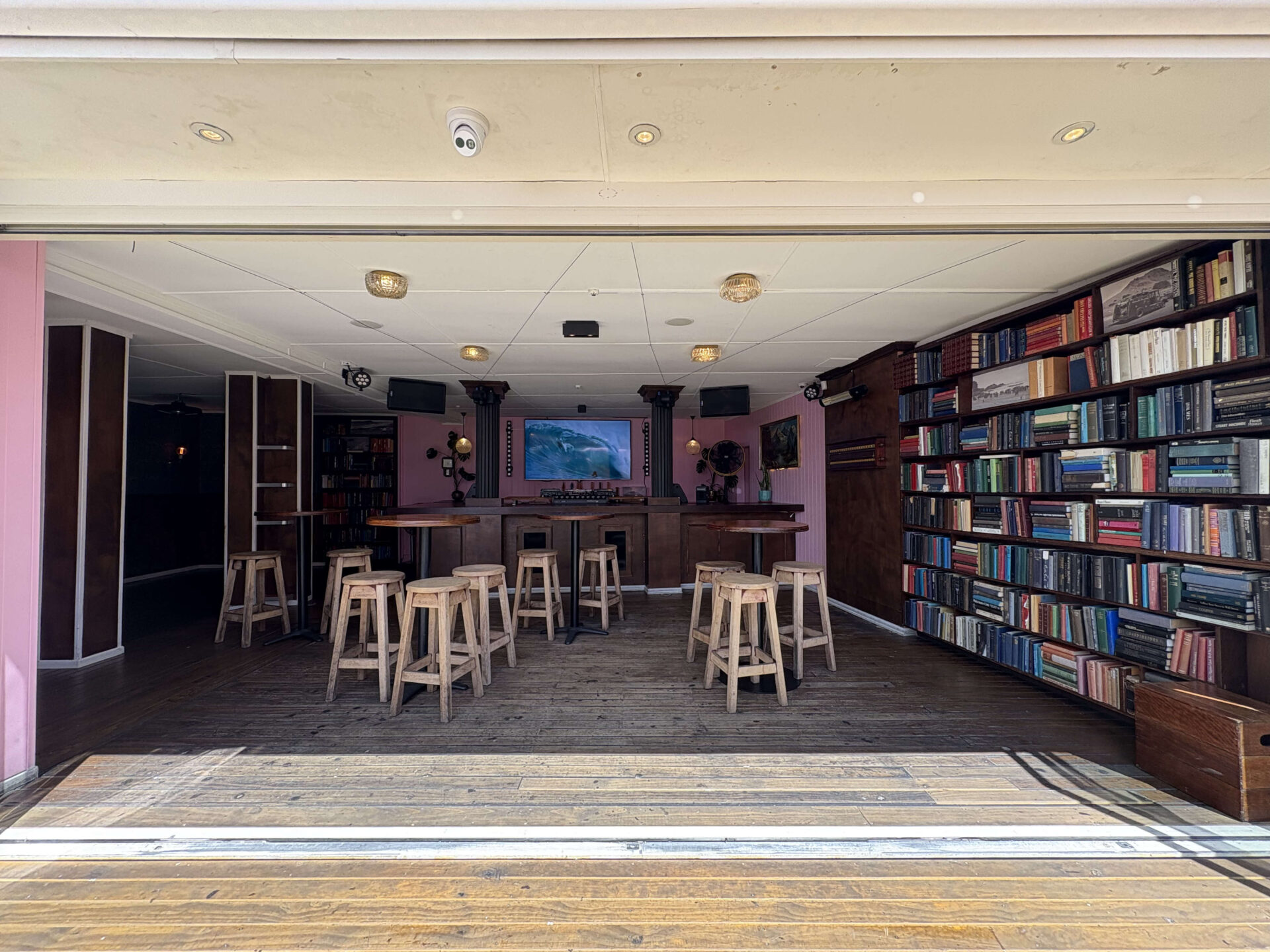 Pink-walled interior room with timber bar, bookshelves floor-to-ceiling, and backless stools on wooden floor