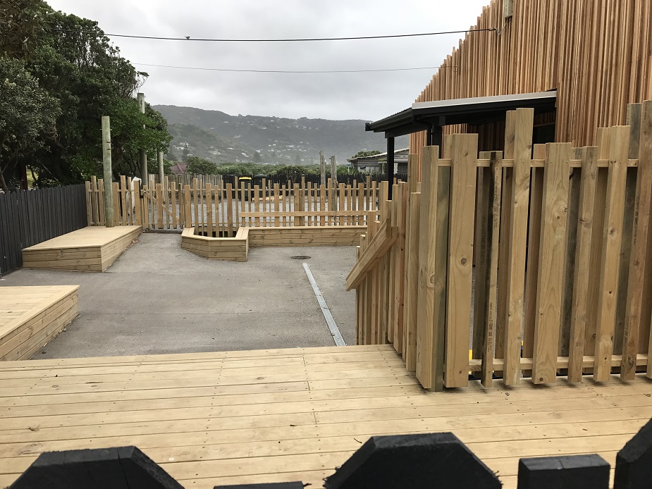 Timber-battened building exterior with open courtyard, low timber seating and fencing, overcast hillside backdrop