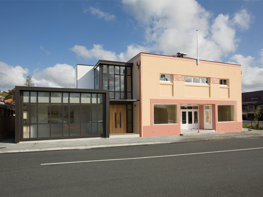Salmon-rendered two-storey hall with glass extension and dark-framed shopfront, street view