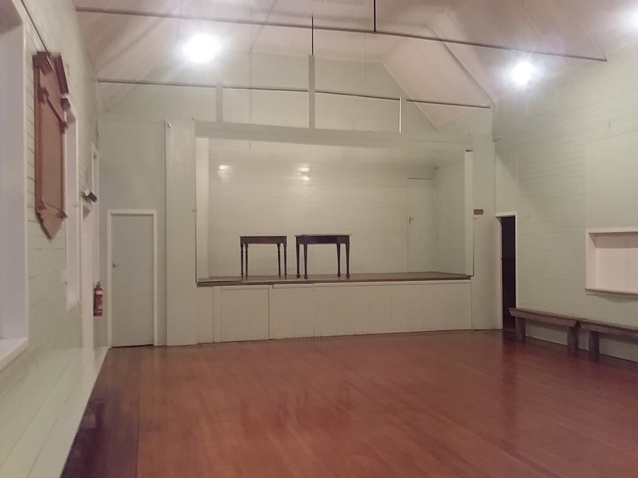 Plain white hall interior with timber floor, raised platform, and exposed rafters