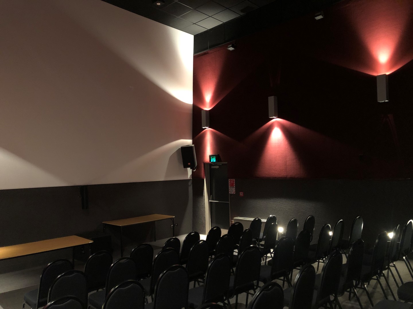 Dark screening room with angled wall panels lit in red, rows of dark chairs facing a blank screen