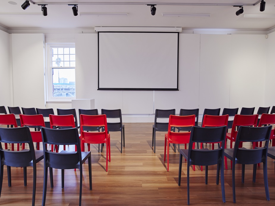 White-walled room with red and dark chairs in rows, projection screen, timber floor