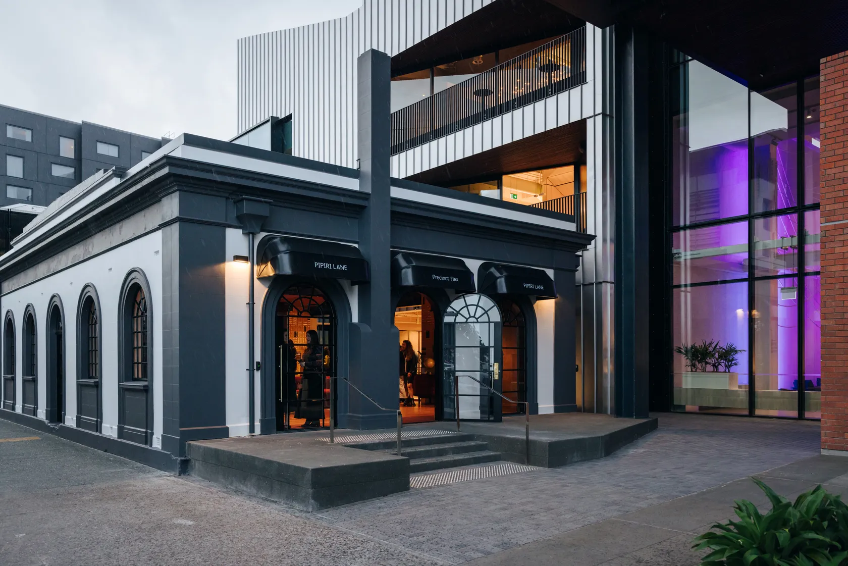 Dark rendered heritage building with arched shopfront openings and modern upper addition, street entrance at dusk