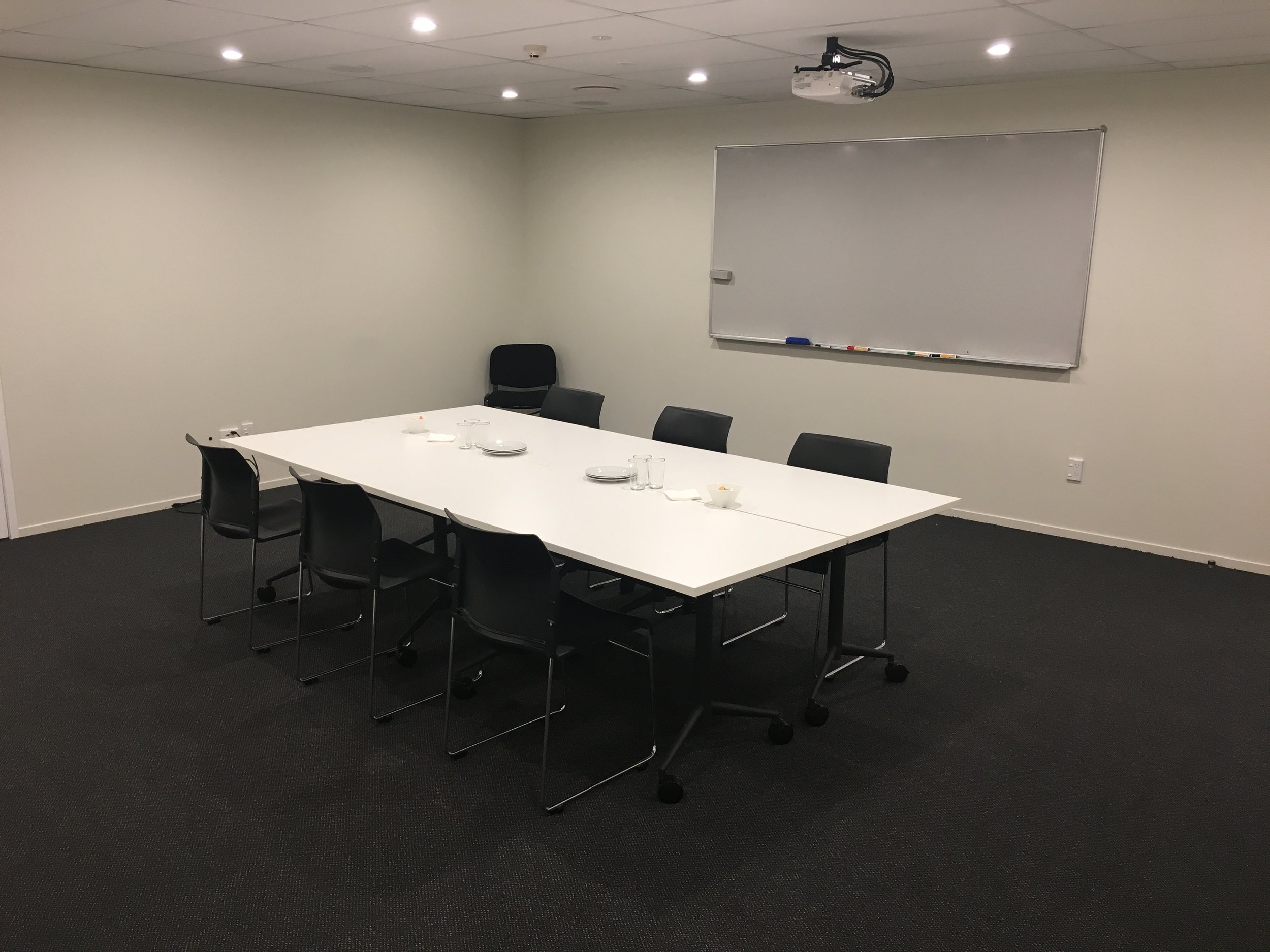 Plain meeting room with a white rectangular table, dark chairs, whiteboard, and projector