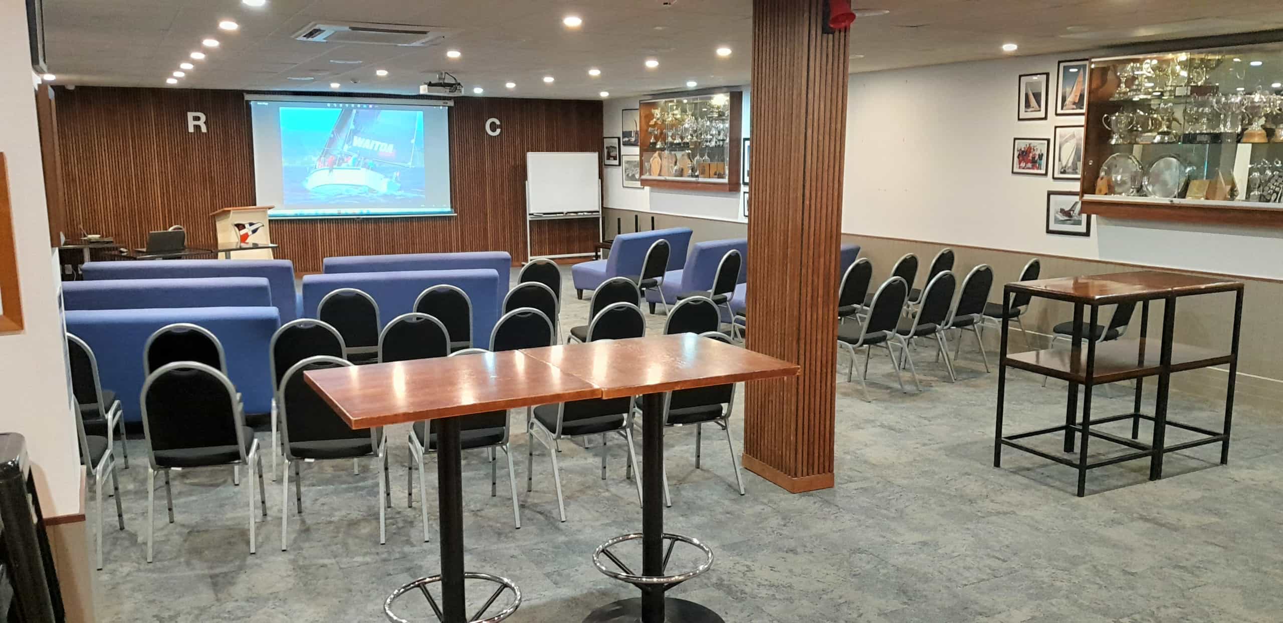 Timber-panelled function room with rows of blue and black chairs facing a projection screen and lectern