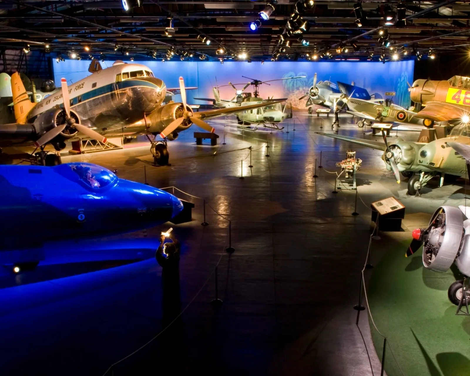 Large darkened museum hall with multiple historic aircraft on display under coloured spotlights