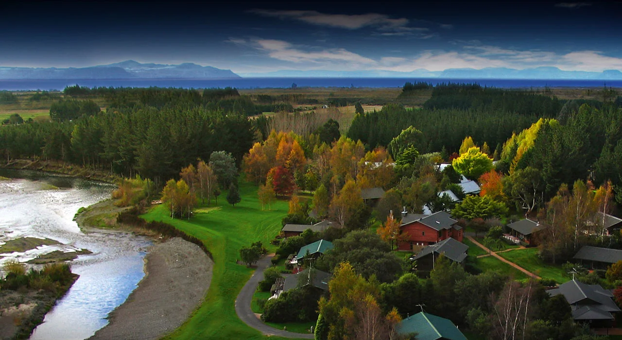 Aerial view of lodge buildings among autumn trees beside a river, lake in background
