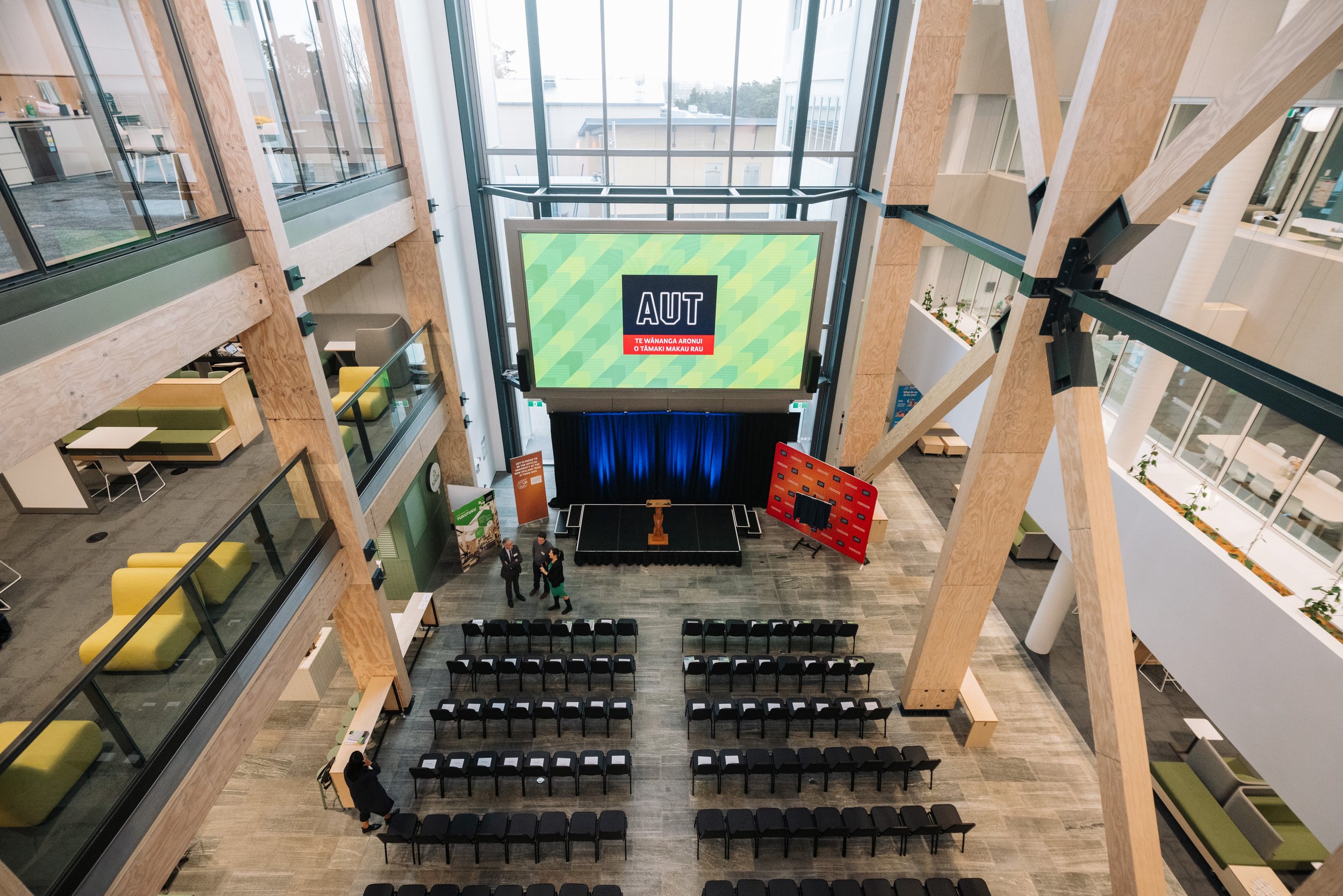 Large atrium event space with timber columns, rows of chairs, and a raised screen displaying a logo