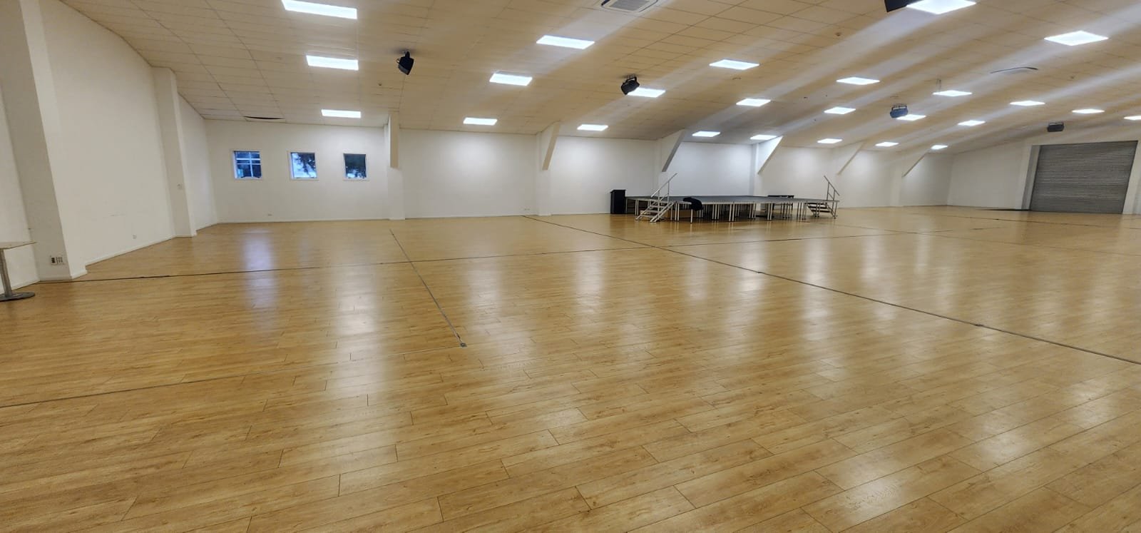 Large empty hall with timber floor and raised platform at the far end, lit by overhead panels