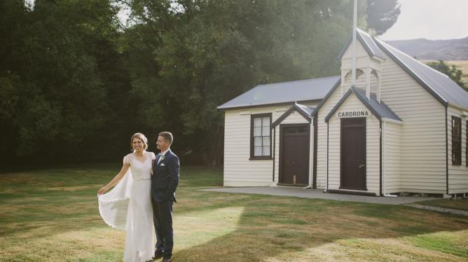 A couple in wedding attire on a lawn beside a small white weatherboard chapel, framed by tall trees with hills behind
