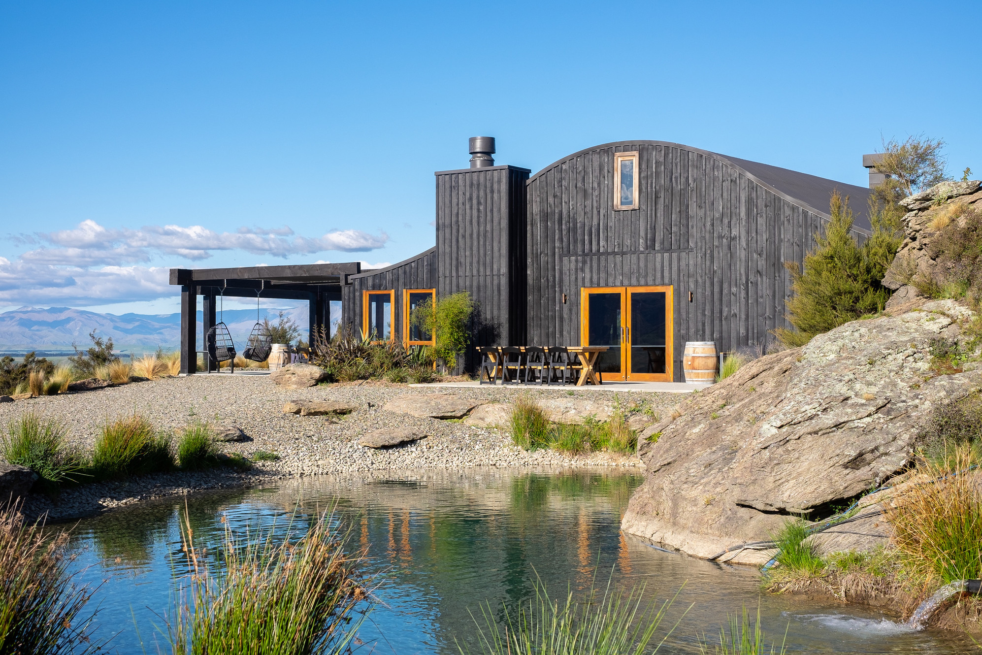 Dark timber barn with curved roof beside a pond, rocky landscape, open mountain backdrop