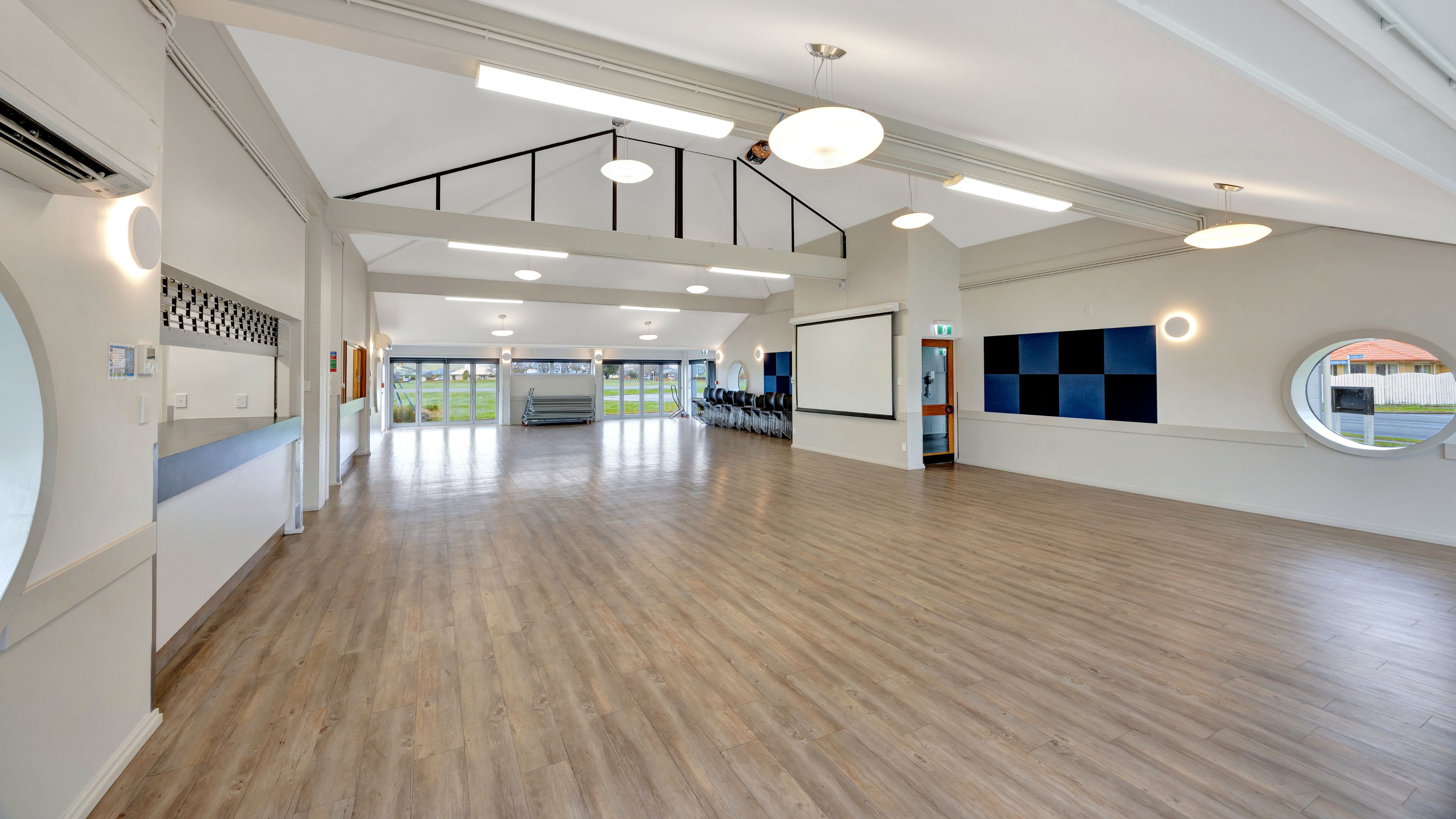 Empty community hall with timber floor, high ceiling, and projection screen
