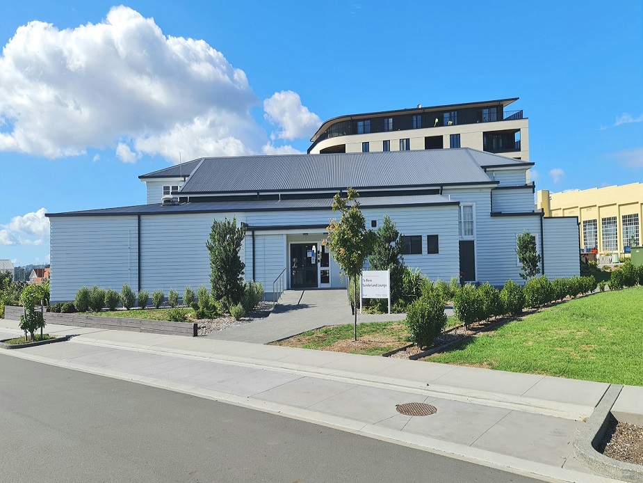 Single-storey white weatherboard building with dark trim, sloped metal roof, suburban setting
