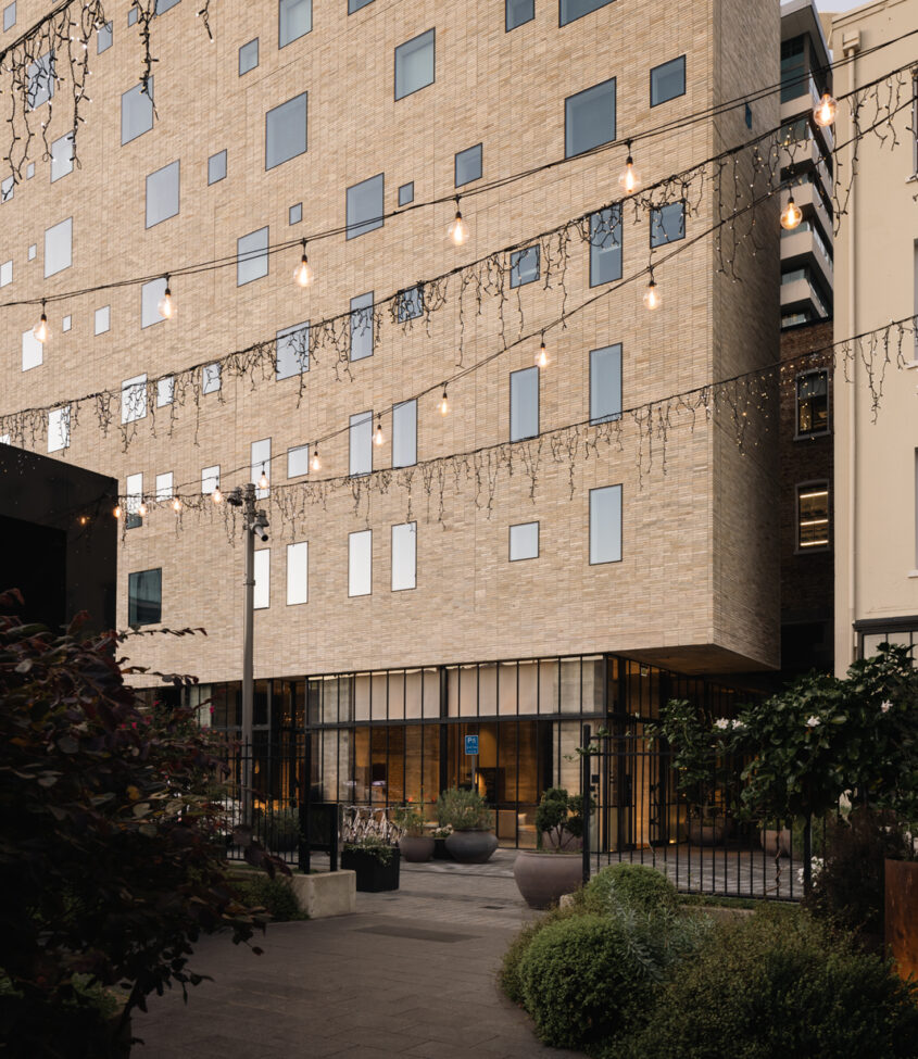 Pale brick building exterior with laneway, string lights overhead and planting at ground level