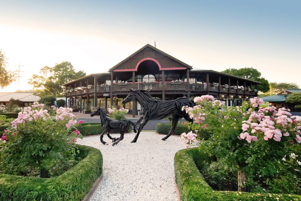 Dark timber two-storey pavilion with a veranda, flanked by formal gardens and horse sculptures