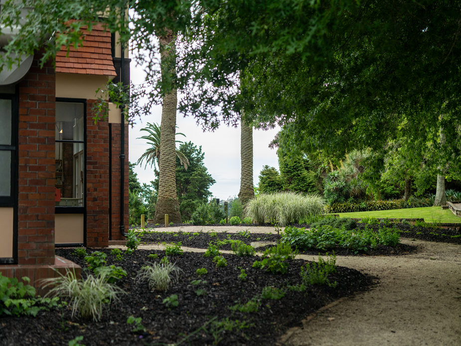 Homestead brick corner with mature planted garden beds, tall palms and trees in background
