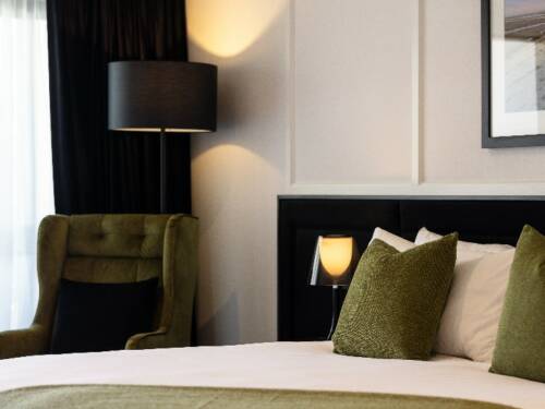 Hotel room with double bed, green velvet armchair, dark bedhead, and bedside lamp