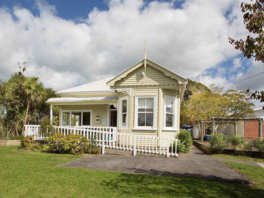 Cream weatherboard villa with verandah, bay window, white picket fence, and lawn, sunny suburban setting