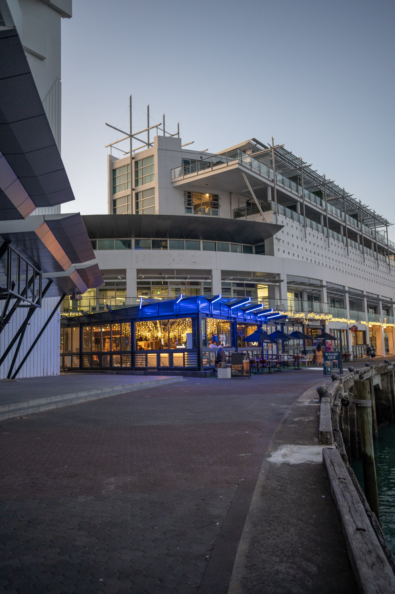 Waterfront venue pavilion with blue neon roof fins and glazed walls, dusk street view