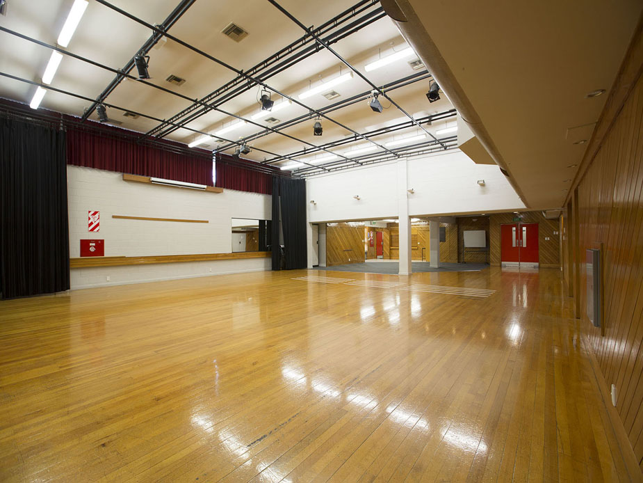 Large hall with polished timber floor, stage lighting rig, and heavy curtains