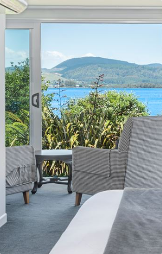 Bedroom interior with armchairs facing large windows overlooking a lake, planting and hills beyond
