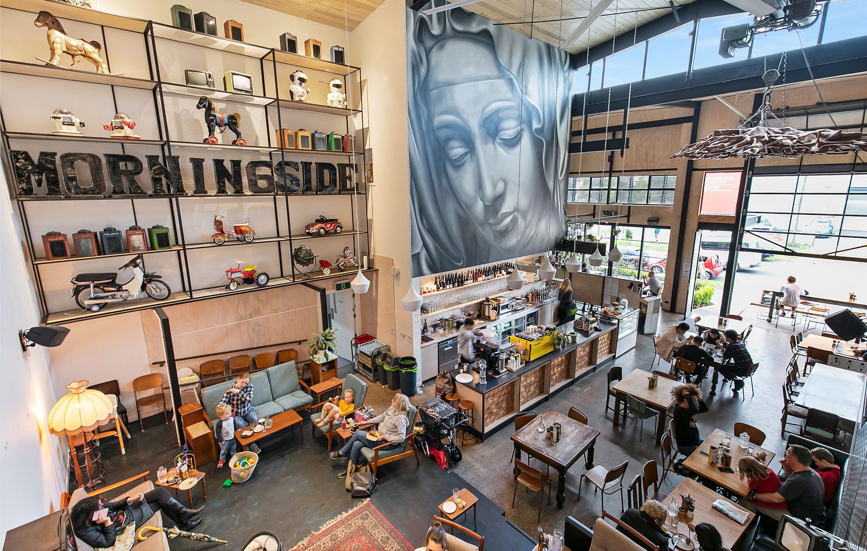 Bird's-eye view of double-height cafe with central bar, large face mural on wall, mixed seating throughout