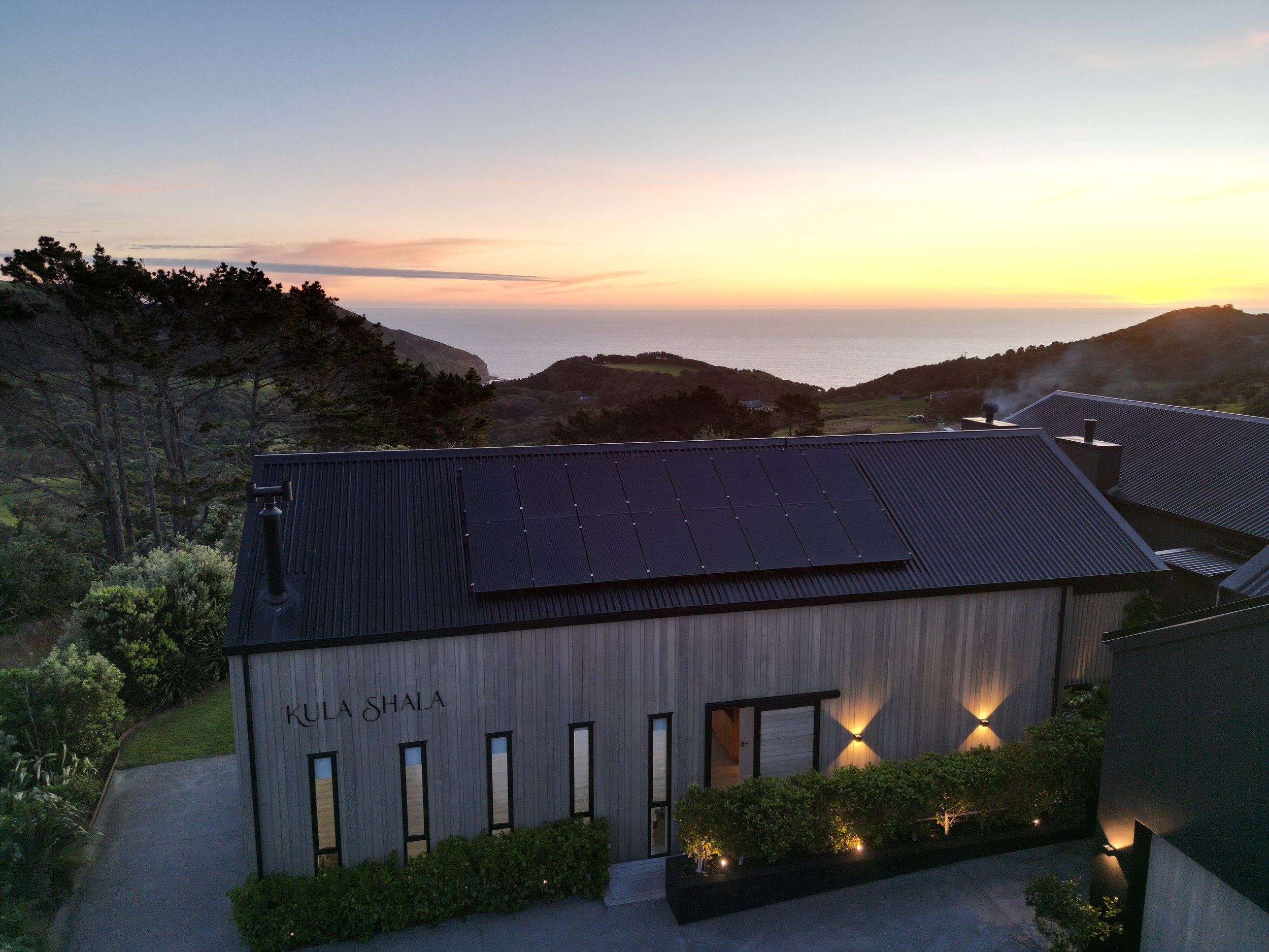 Timber-clad building with a dark metal roof and solar panels, viewed from above at sunset with sea behind