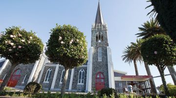 Dark basalt stone church with tall spire, pointed arched windows, and trees in the forecourt