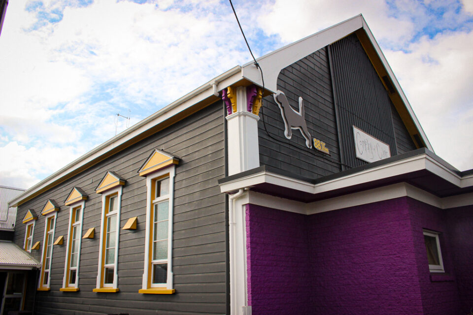 Grey weatherboard building with yellow-trimmed windows and purple brick lower section, street exterior