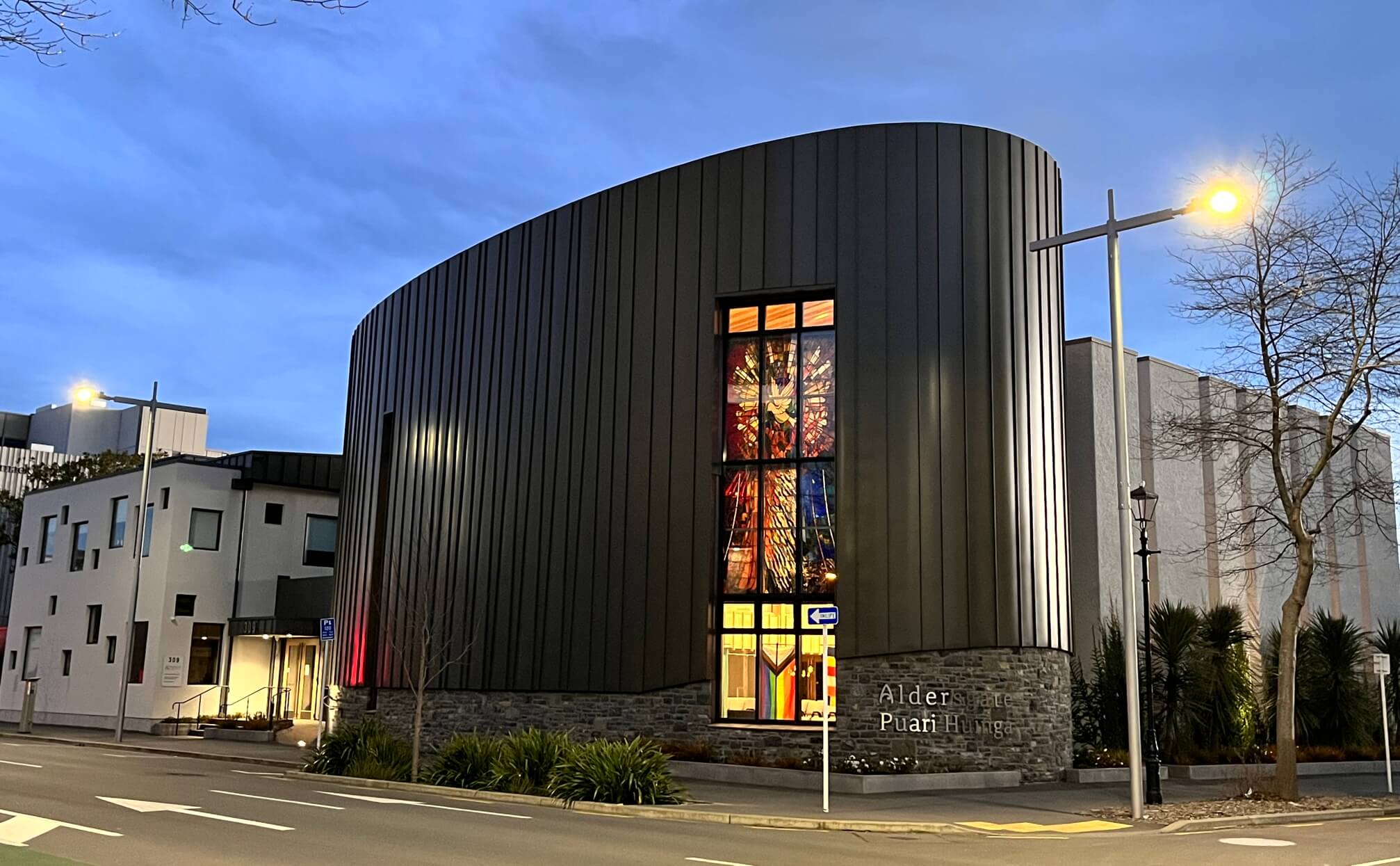 Contemporary curved dark-clad church building at dusk with stained glass feature window