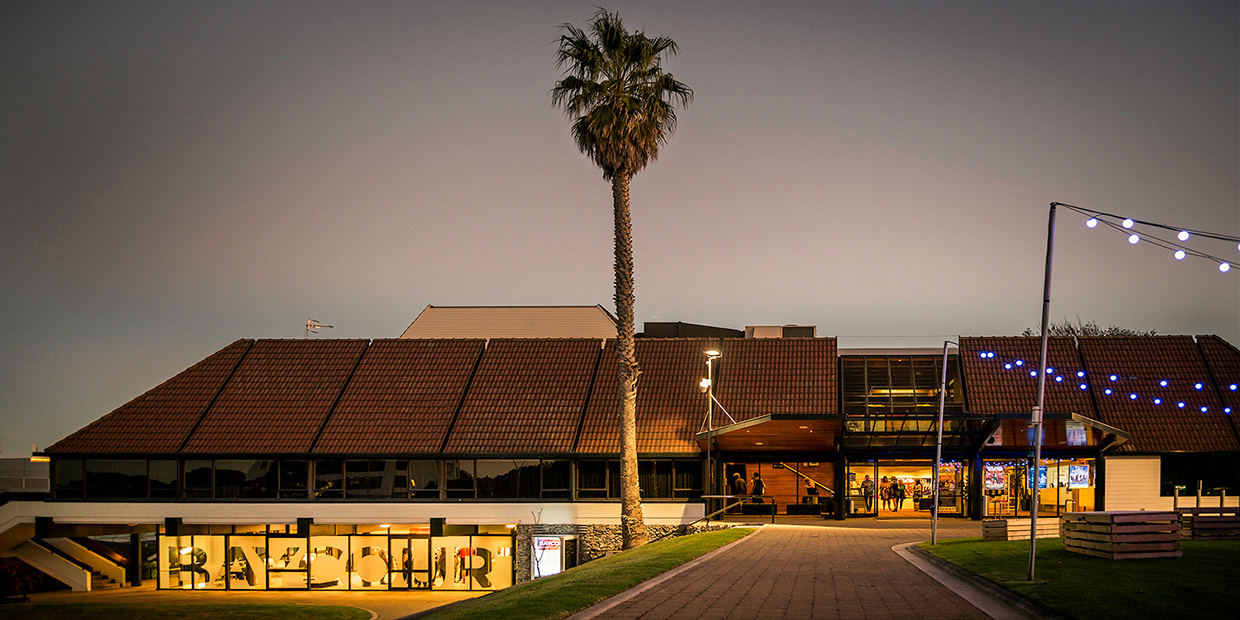 Low-rise arts centre exterior at dusk with a tall palm and festoon lights along the facade