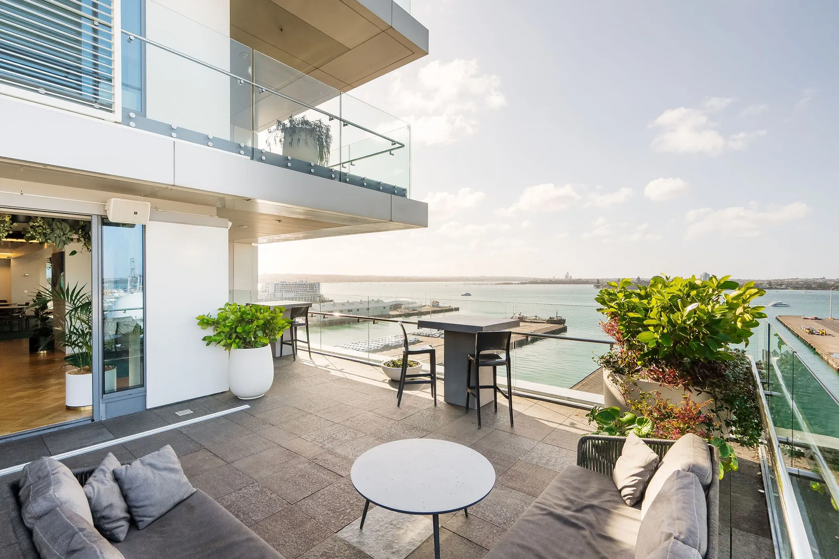 Rooftop terrace with grey sofas and planters, glass balustrade, and harbour and cityscape view