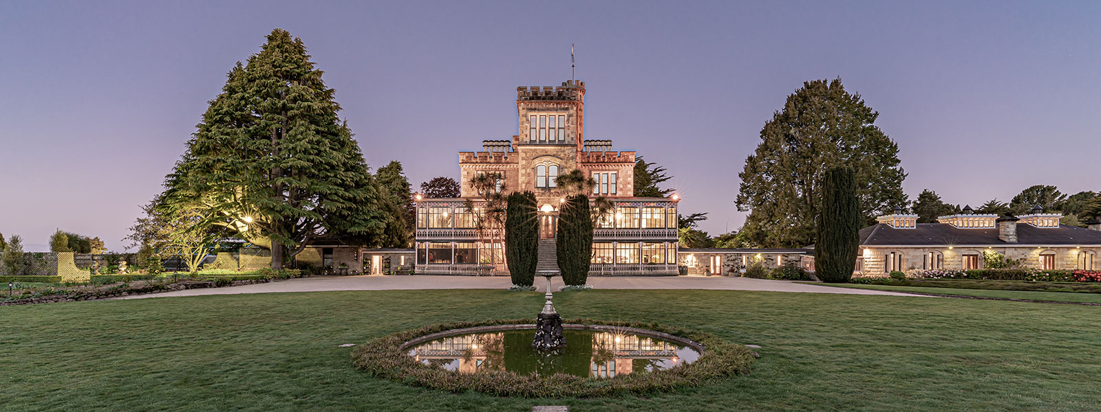 A lit stone castle with a tower and glasshouse wing at dusk, framed by tall trees and a formal lawn with a circular fountain pool