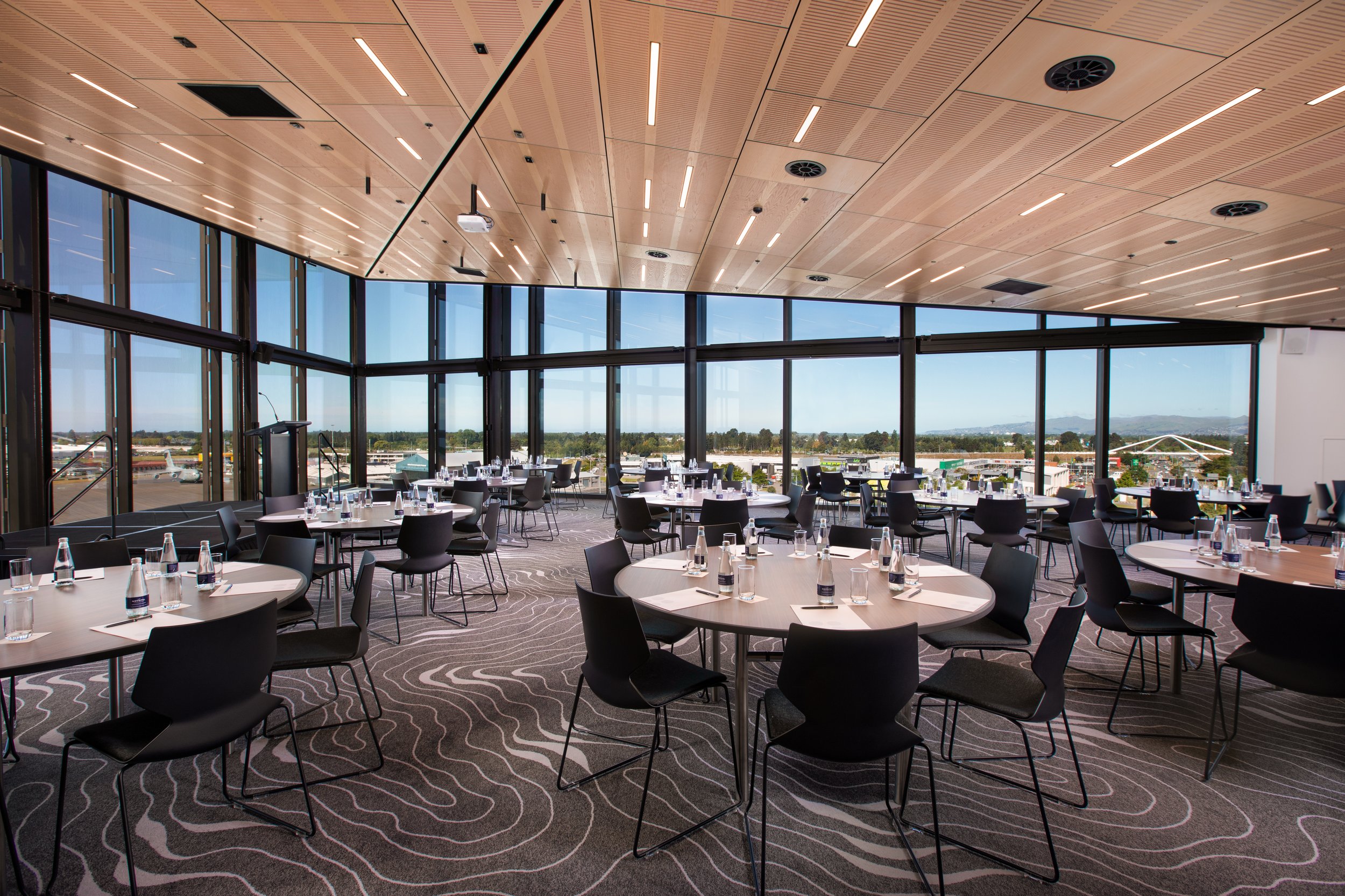 Light timber ceiling event room with round tables and black chairs, floor-to-ceiling windows and open views