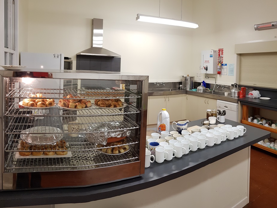 Hall kitchen with stainless benches, display warmer with baked goods, and mugs lined up on counter